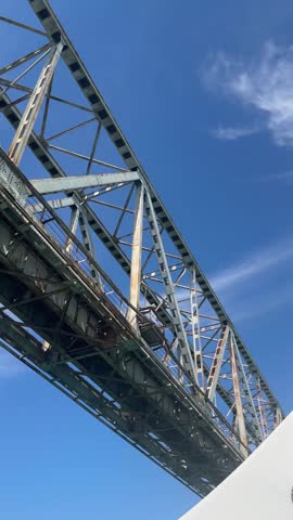 Vertical view of a large bridge passing above a boat on the water. The footage captures architectural structure, blue summer sky and the perspective of traveling by boat beneath a bridge, suitable for travel, transportation, tourism and boating concepts.