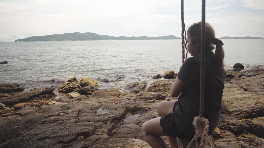 Woman sits on a rustic rope swing by the sea, sad and lost in thought as she gazes out over the rocky shoreline and calm ocean waters of Khao Laem Ya–Mu Ko Samet National Park