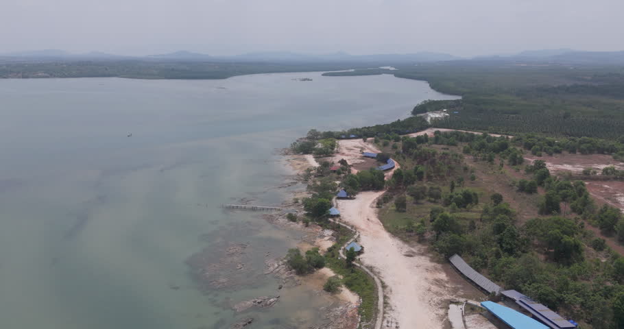 Backward aerial view of a scenic coastal landscape with a winding dirt path, wooden piers, and lush greenery along the shore.