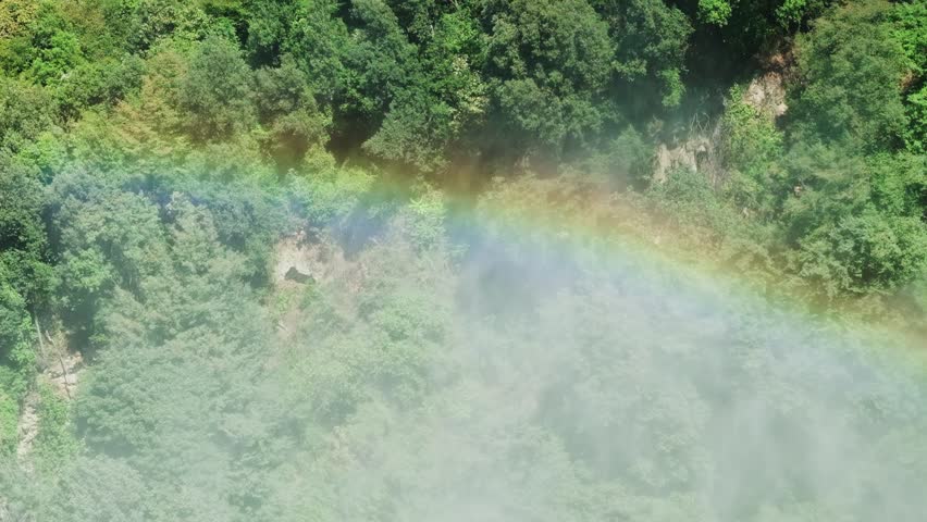 Overhead view of a vivid rainbow stretching across a verdant green valley, with gentle mist rising. The slow-motion perspective emphasizes the tranquil and breathtaking natural environment.