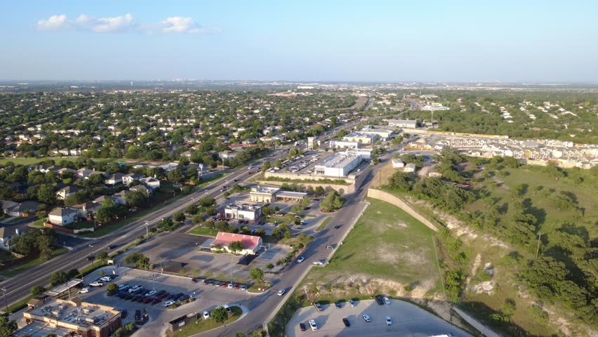Elevated drone perspective reveals active parking lots, gas station bays, fast food drive thru along Potranco Road. Arterial traffic flows smoothly under late afternoon sunlight. San Antonio, TX. USA