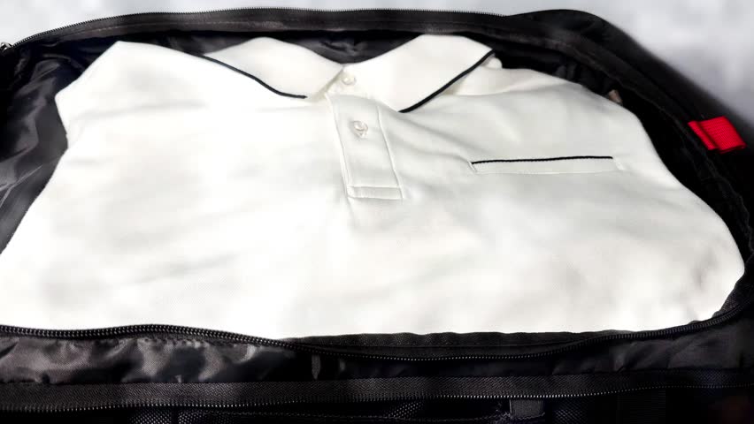 Closeup of a man’s hands packing a travel case, by placing a bottle of aftershave lotion and a cordless electric shaver on top of a clean white casual shirt.
