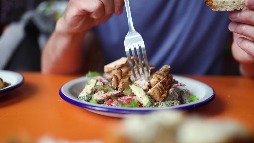Enjoying a fresh salad with grilled chicken and cheese on a vibrant table in a restaurant setting during lunch time. Person eating fresh chicken caesar salad with a fork.