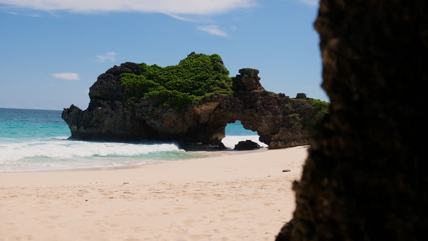 Large ocean waves crash onto a white sandy tropical beach beneath a dramatic natural rock arch on the coast of Sumba, Indonesia. View of turquoise water, breaking waves and a secluded tropical shoreline.
