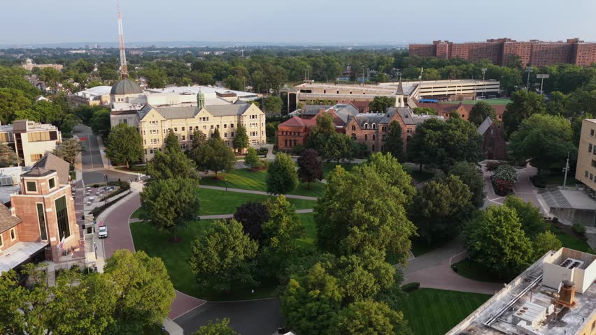 Seton Hall University campus quad in South Orange, New Jersey showcases historic brick buildings, a domed clock tower and manicured lawns amid dense green summer trees. Aerial drone view.