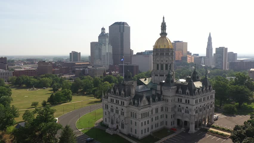 Cinematic aerial footage captures Connecticut state capitol building in Hartford featuring golden dome architecture surrounded by urban park landscape under clear bright summer sky.