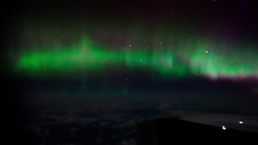 Breathtaking aerial footage captures mesmerizing green and purple northern lights dancing across dark night sky above thick cloud layer as seen from passenger aircraft window.
