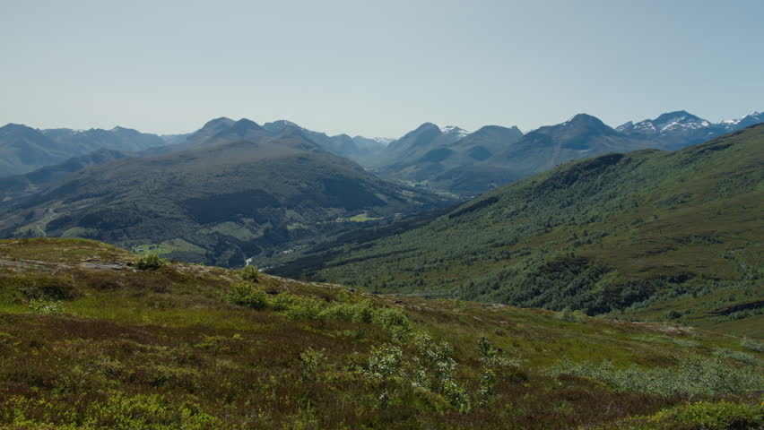 Heather-covered highland ground looks across a deep green valley toward distant Sunnmøre peaks in bright midday light.