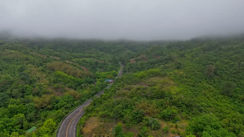 Aerial drone footage captured over winding asphalt road curving through vibrant green tropical hillside mountains with low hanging clouds mist and fog in tropical landscape setting.
