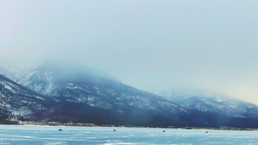 Cars driving on frozen Baikal lake under mountain range background. Winter Siberia landscape view, ice surface, snow covered peaks in fog. Travel adventure, outdoor exploration and extreme hobby.