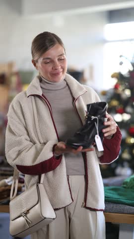Adult woman in a jacket is choosing stylish ankle boots with fur against the background of a Christmas tree in a clothing and shoe store. Buyer came for shoes at bargain prices. High quality 4k