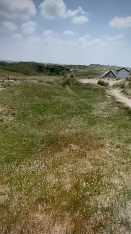 Colorful beach cabins dot the sandy dunes at Gouville-sur-Mer, Normandy, France. Each cabin features a vibrant, painted roof, standing amidst grassy dunes. The ocean stretches out to the right, its turquoise water contrasting with the sand and green grass. The sky is partly cloudy, adding a picturesque backdrop to this serene coastal landscape.