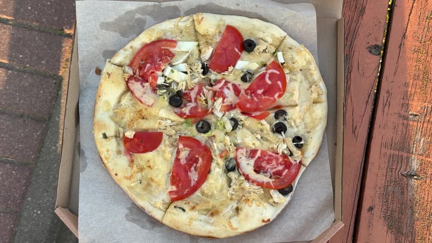 A sliced pizza with tomatoes, black olives and chicken sits on paper in a cardboard box. Fast food, takeaway meal, Italian cuisine, outdoor lunch.