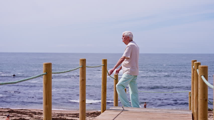Porto. Portugal. An elegant senior man standing on a wooden pier, pensively looking at the ocean waves on a sunny day. He is enjoying his retirement