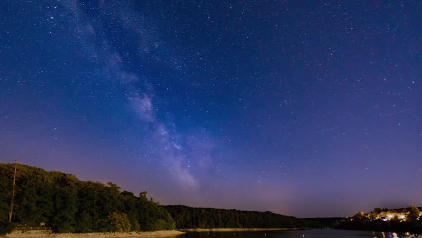 A stunning nighttime time lapse with a slow camera zoom in showing the Milky Way galaxy moving across the starry sky above a peaceful river surrounded by silhouettes of trees.
