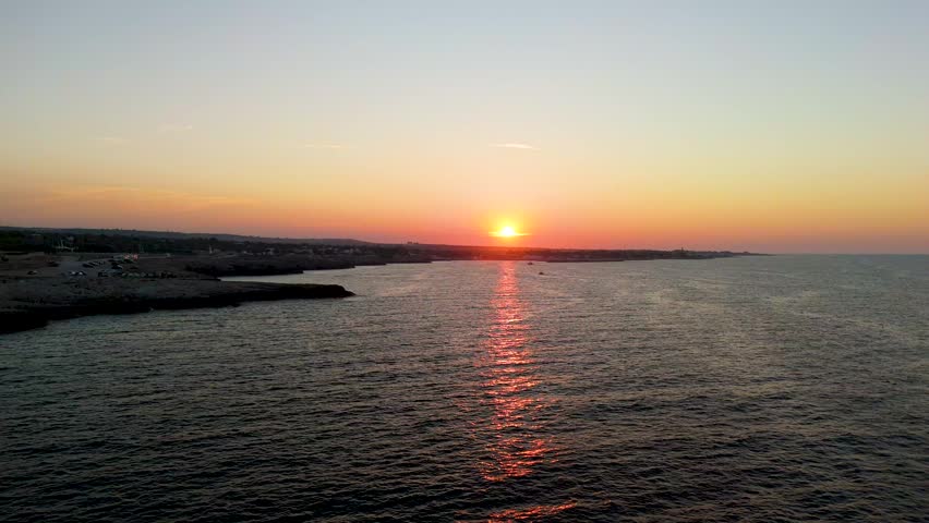 Sun setting above dark coastline at Polignano a Mare with red light reflections shimmering on calm sea surface