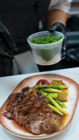 Chef garnishing grilled steak with chopped chives. Green vegetables and a browned potato portion sit on the ceramic plate.