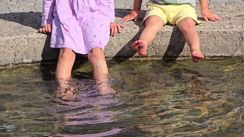 An editorial stock photo capturing children sitting on the stone ledge of a public fountain, splashing and dipping their feet into the water on a warm summer day. The composition focuses on their legs, water ripples, and stone paving. This image is ideal for editorial projects, articles on family lifestyle, summer vacations, and city life.