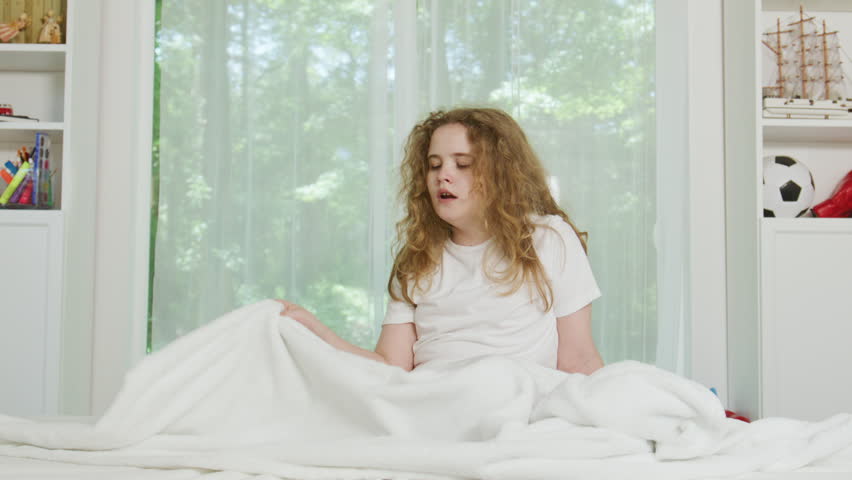 Child wakes while sitting in bed. Child looks tired during morning routine. Sleepy child rests beneath white blanket. Young child yawns beside bedroom window. Waking up kid a quiet morning.