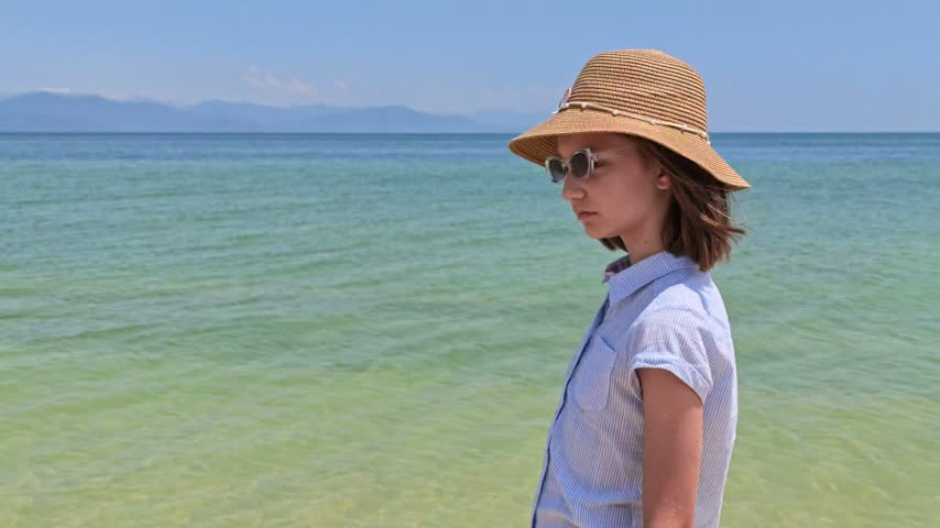 Child girl in straw hat stands near calm ocean. She looks out at distant mountains under clear sky