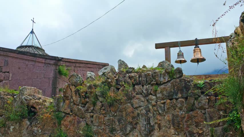 Ancient stone wall with hanging bells. Red brick church with domed roof. Mountainous backdrop under cloudy sky