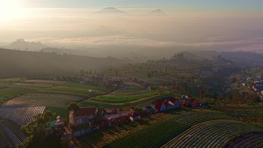 Sunrise aerial view over agricultural terraces, misty clouds rolling through a mountain valley, rural village and lush green fields from above.