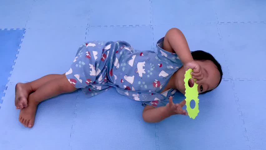 A young child is lying on a blue floor mat while playing with a small toy in a home environment.