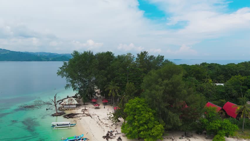 Red tile rooftops and tall palm trees fill small coastal village near Gili Nanggu, Lombok, Indonesia, green mosque dome rises among misty mountain ridges in distance. Aerial drone rising shot