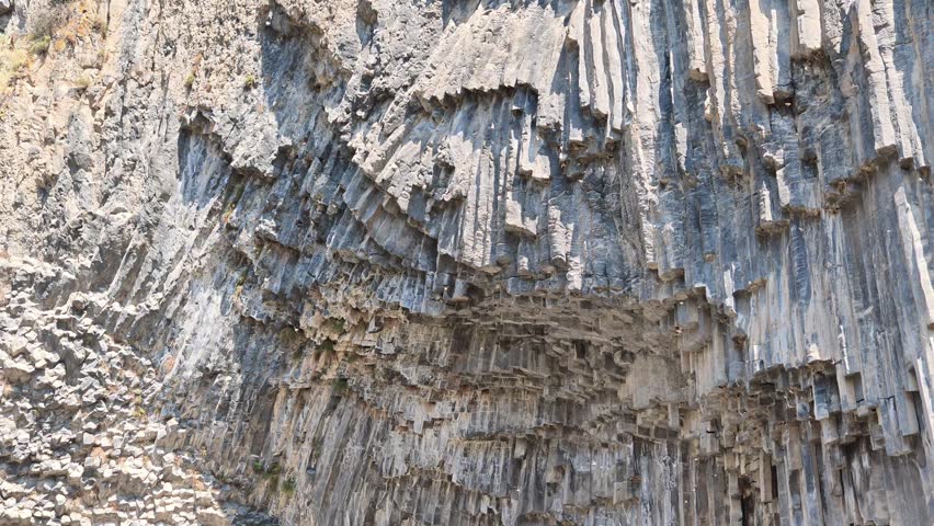 Rugged basalt columns rise vertically in layered formation. Sunlight highlights textured stone surfaces. Natural erosion shapes intricate geological patterns. Rock faces display weathered