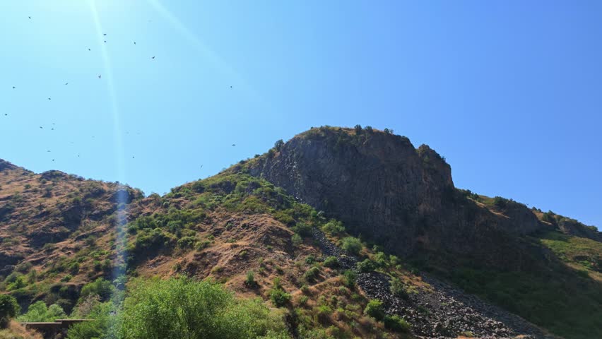 Sunlight glares over rugged mountain ridge. Green shrubs dot dry slopes under clear blue sky. Birds fly above rocky terrain