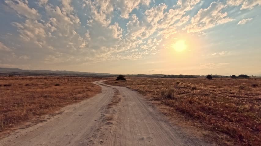 Sunset over dirt road through dry grassland. Sky features scattered clouds. Road curves gently into distance. Single bush stands near center. Horizon shows rolling hills