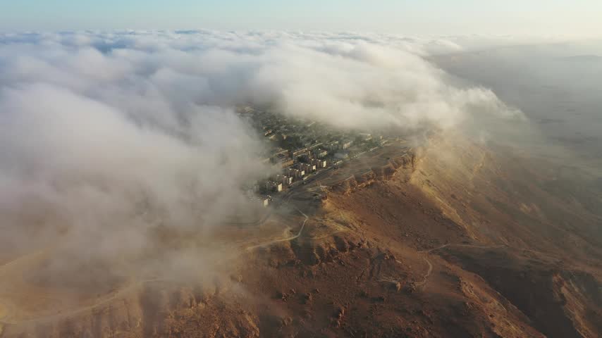 Aerial view of Mitzpe Ramon town perched on the edge of the Makhtesh Ramon crater with low clouds and golden hour light over the desert landscape in Mitzpe Ramon, South District, Israel.