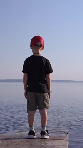 Little boy standing on the edge of a concrete pier, looking thoughtfully at the calm expanse of water. The child contemplates the serene landscape during a peaceful summer day by the lake