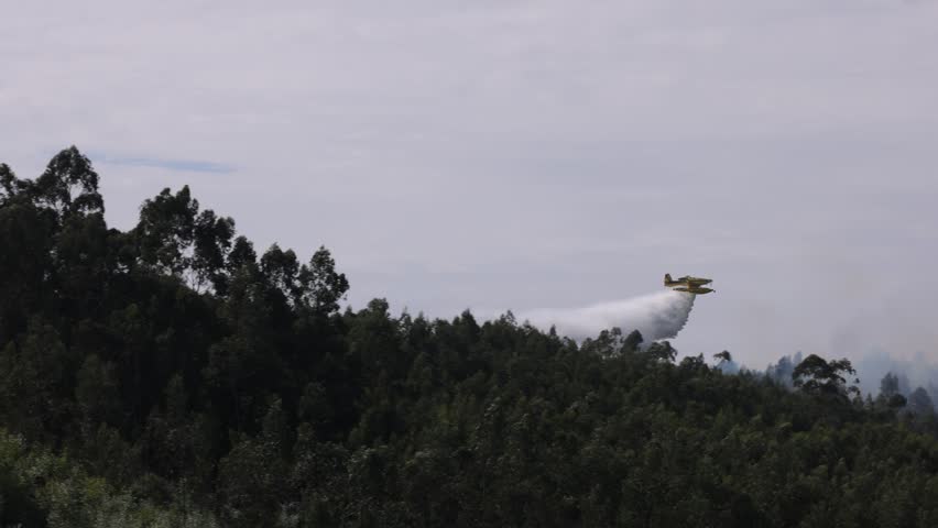 Dramatic aerial footage in slow motion shows a fire fighting airplane releasing a huge water drop over a smoky forest wildfire during a heatwave in Portugal, with morning light and thick haze.