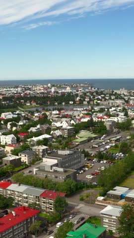 An aerial view shows the city of Reykjavik, Iceland on a sunny day. The cityscape extends to the North Atlantic Ocean, with a small lake and colorful buildings visible.