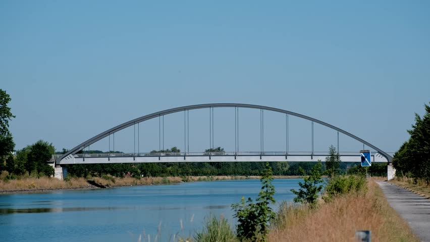 A beautiful architectural view of a tied-arch bridge crossing a peaceful shipping canal under a clear blue sky.