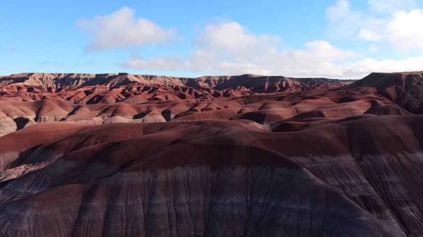 Aerial view of the colorful Painted Desert badlands with rolling hills and distant clouds under a blue sky in Winslow, Arizona, United States.