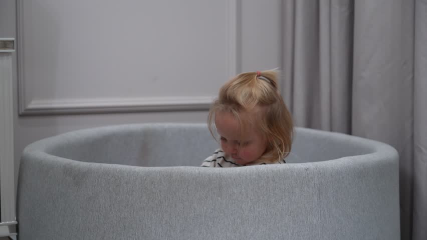 Cute toddler climbs out of padded playpen filled with soft balls, illustrating childhood exploration and growth.