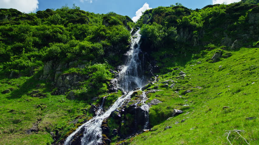 Scenic mountain waterfall cascades over dark rocks through lush green alpine vegetation along Romania Transfagarasan route in summer.