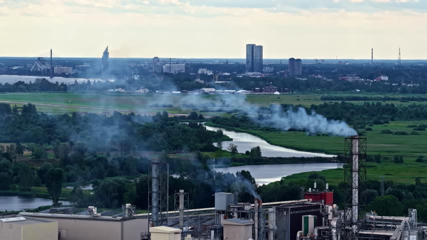 Aerial view of an industrial plant with smoking chimneys, lush green fields, and a winding river, with the city skyline visible in the distance in Latvia.