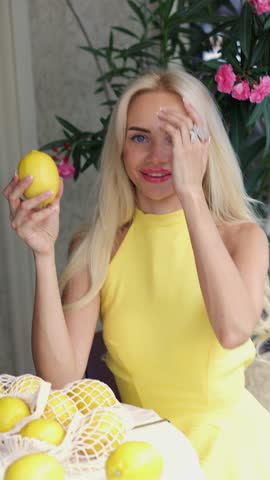 "Vertical medium slow motion video of a gorgeous young Caucasian blonde woman with blue eyes, holding a fresh whole lemon near her face and smiling dreamily. She turns her head, looks at the camera, and poses with a string bag full of lemons at a table. Bright summer light, green plants with pink flowers background. Concept of healthy lifestyle, citrus fruits, vitamin C, organic skincare, natural beauty, and summer vibes."