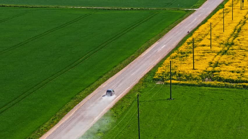 Aerial view of a car driving along a dusty dirt road through vibrant green and yellow rapeseed fields in the countryside of Latvia.