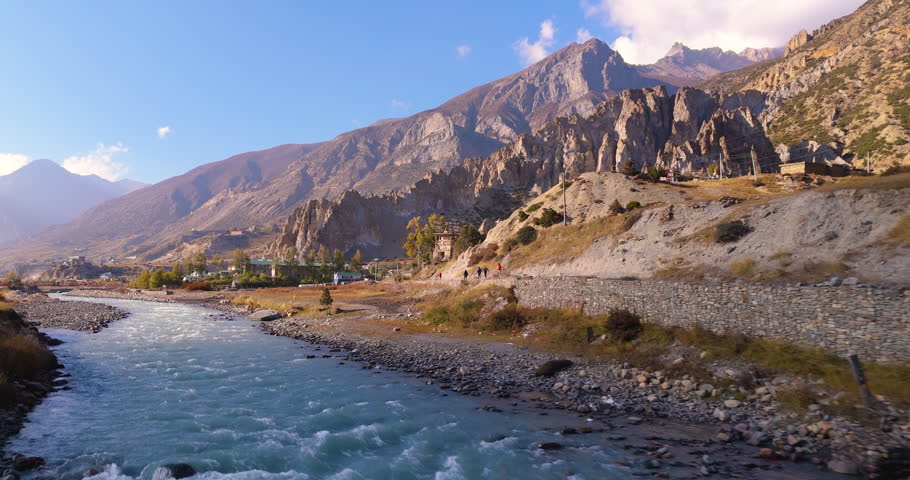 Annapurna Circuit, Manang, Nepal, aerial gliding shot along the Marsyangdi River showing hikers on a trail beneath rocky cliffs near Bhraka Monastery in the Annapurna region.