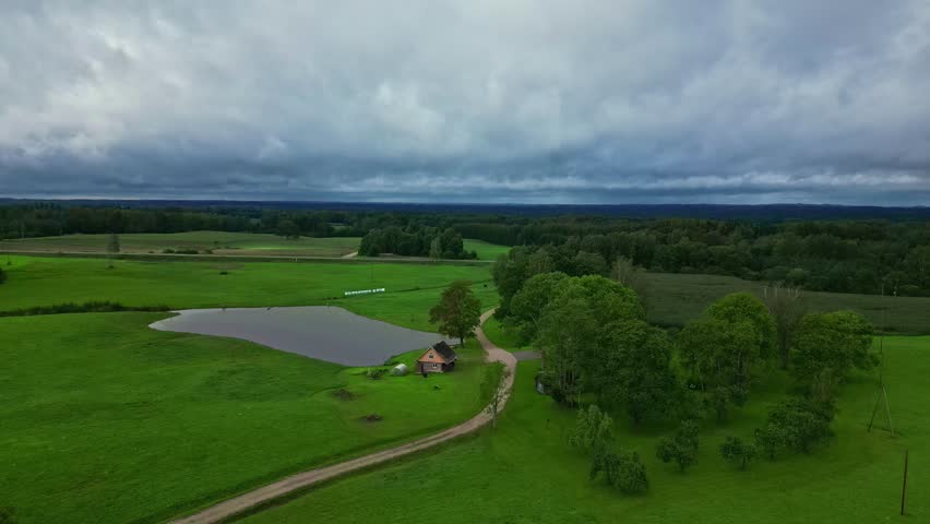 Aerial view of a small house situated next to a pond in a lush green rural landscape under a cloudy sky in Latvia.