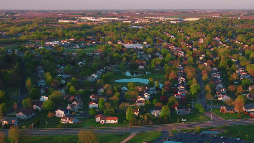 Aerial of single-family homes and suburban rooftops at golden hour in DeForest, Wisconsin, a residential real estate neighborhood with curving streets and green yards.