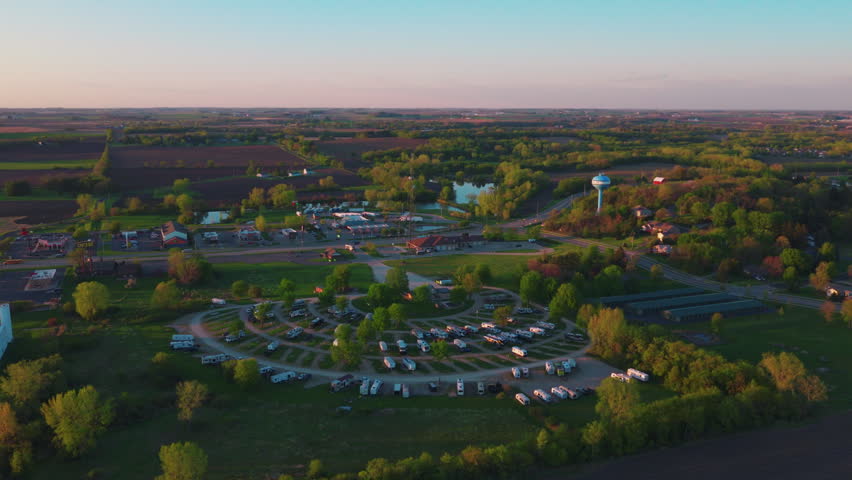 Golden hour drone view of a rural campground near DeForest, Wisconsin, rows of RVs on a circular loop surrounded by farmland, trees and a small pond at sunset.