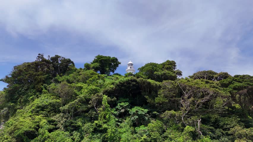 A powerful 4K drone shot looking up at the Kannonzaki Lighthouse amidst lush green trees, set against a dynamic, slightly cloudy sky.

