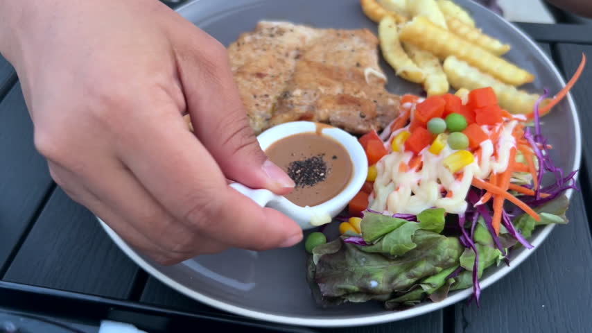 Pouring creamy sauce over fresh salad with grilled pork steak and French fries on plate