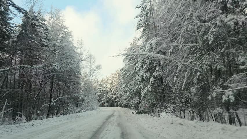Vehicle traveling along a winding snow covered forest road past a warning sign, showing a winter journey through a serene woodland landscape