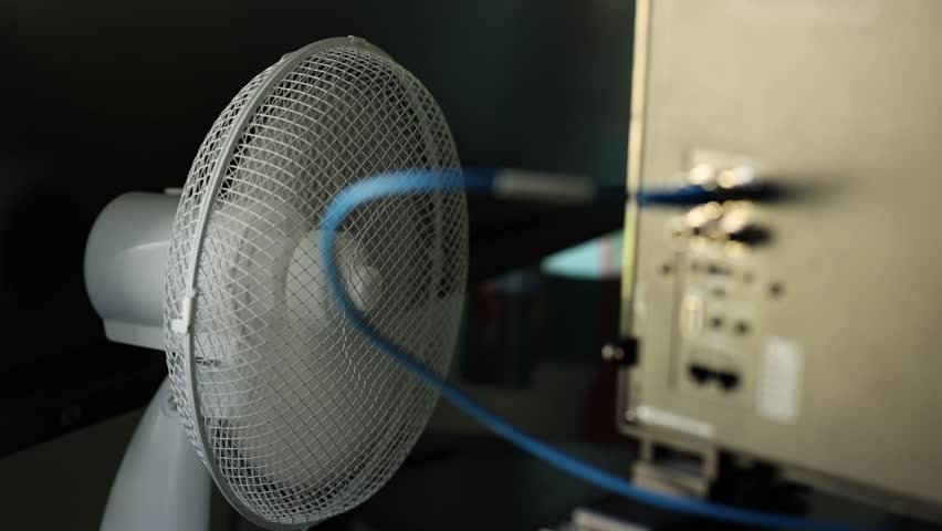 Slow motion of a white desk fan blowing air near computer monitors and electronic server equipment, with a blurred blue cable running past the foreground.